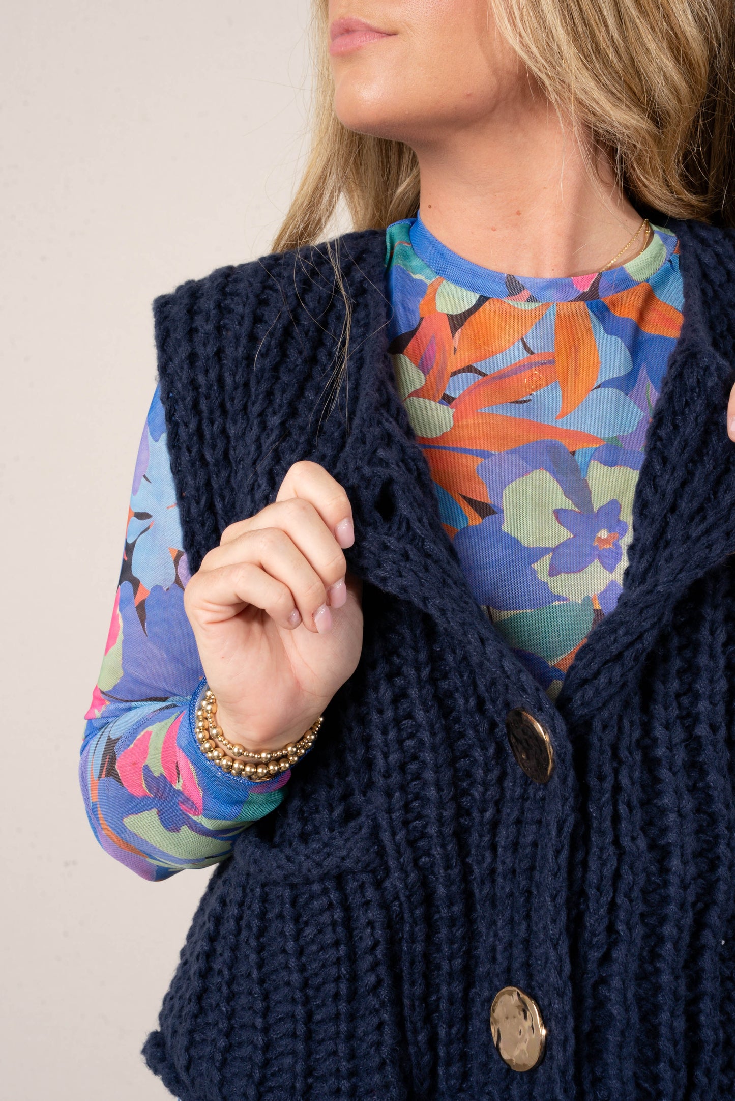 Close up of a woman wearing a bright blue floral print mesh top layered under a navy, chunky knit sweater vest with large gold buttons.