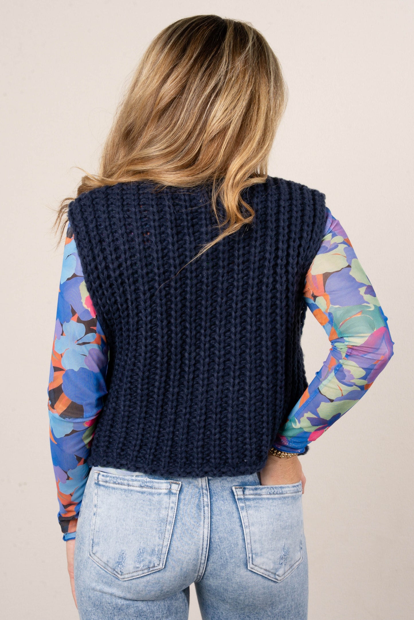 Back of a woman wearing a colorful blue floral mesh long sleeve top layered under a chunky navy knit vest, paired with light wash jeans.