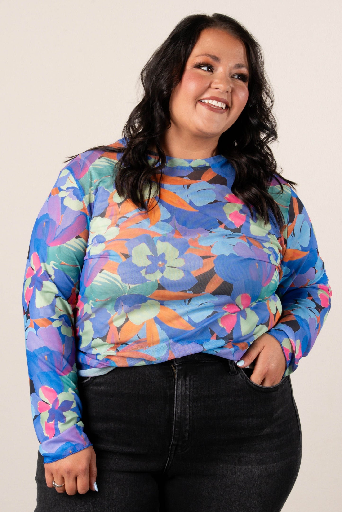 Plus size, smiling woman wearing a colorful floral mesh long sleeve top in blue, orange, pink, and green tones, paired with black high-waisted jeans and casual sneakers.