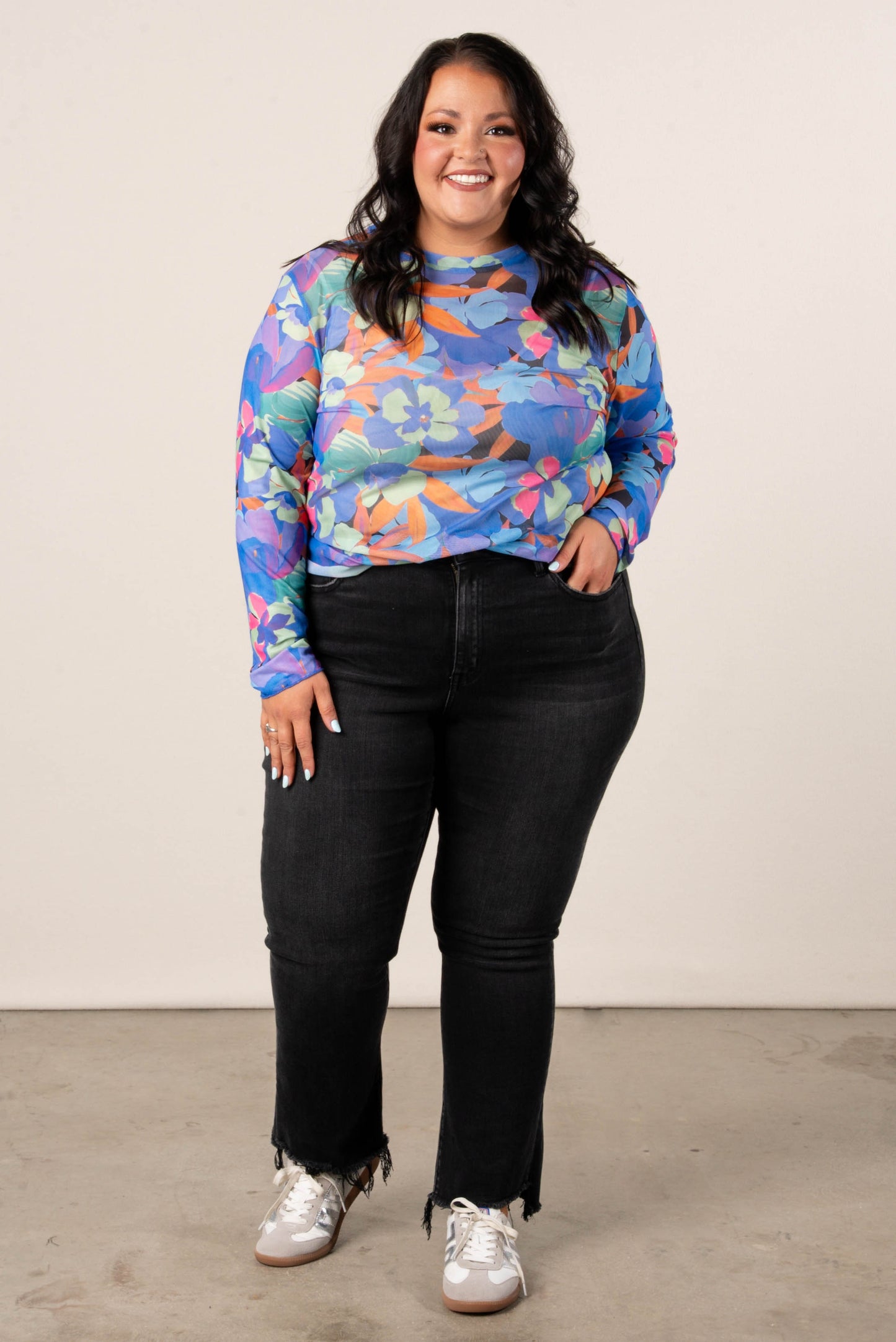 Plus size woman wearing a colorful floral mesh long sleeve top in blue, orange, pink, and green tones, paired with black high-waisted jeans and casual sneakers.