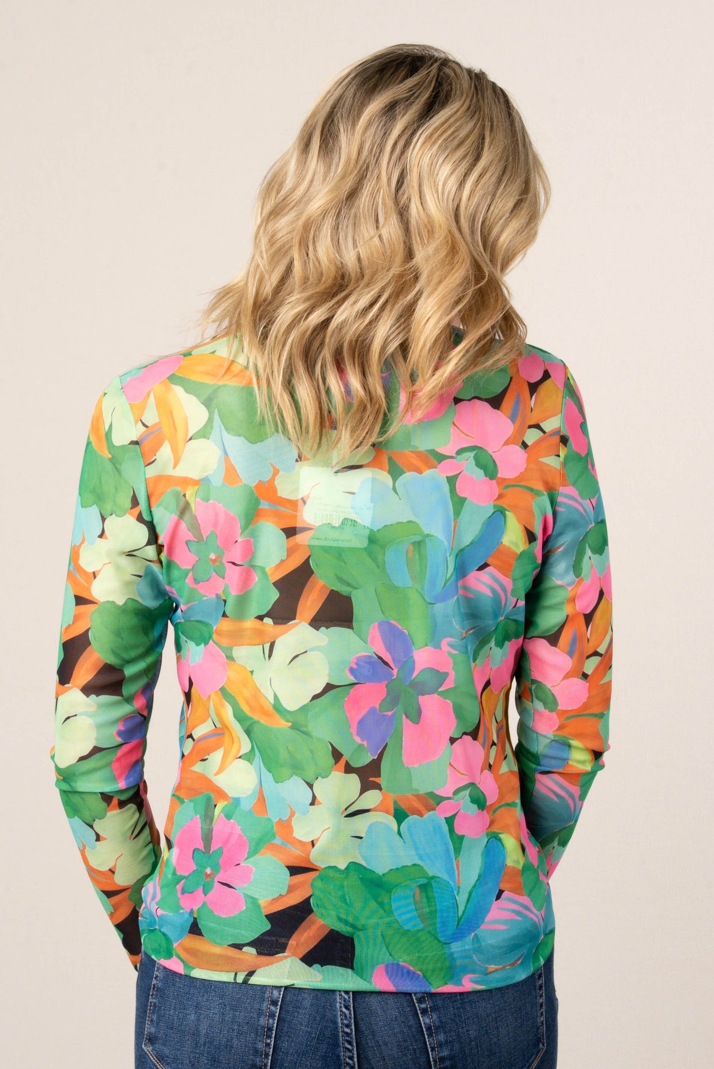 Back of a woman wearing a colorful floral mesh long sleeve top in green, pink, and orange tones, styled with high-waisted blue jeans.