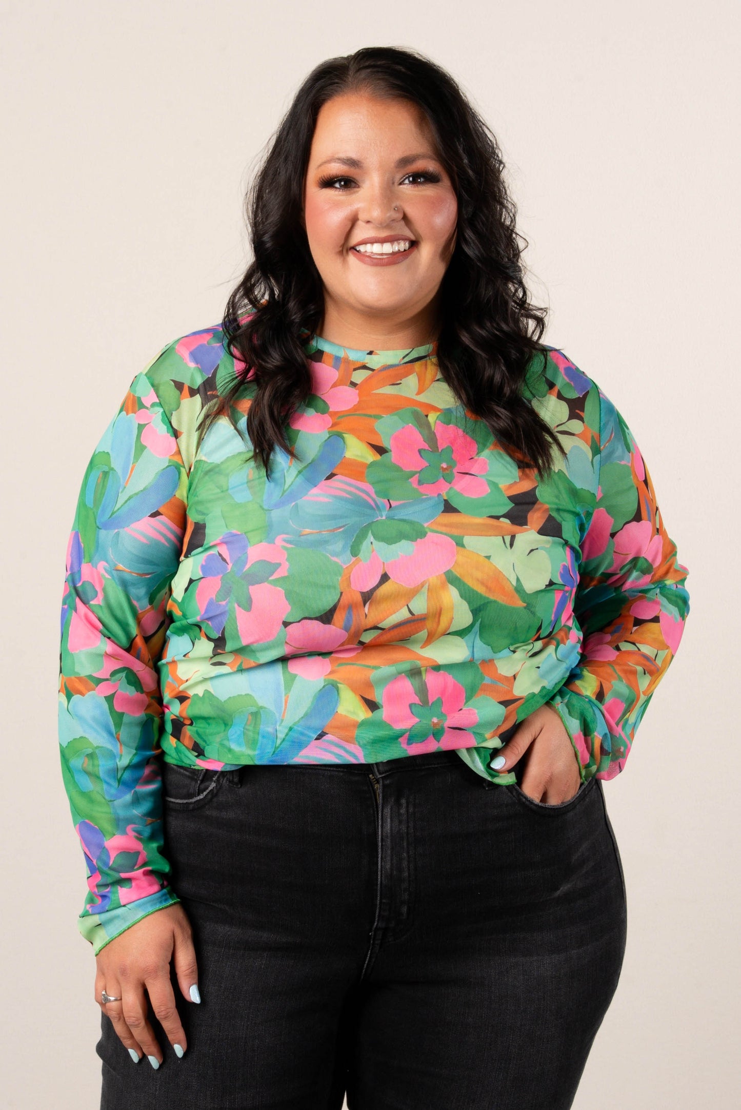 Smiling, plus size woman wearing a colorful floral mesh long sleeve top in green, pink, and orange tones, styled with high-waisted black jeans.