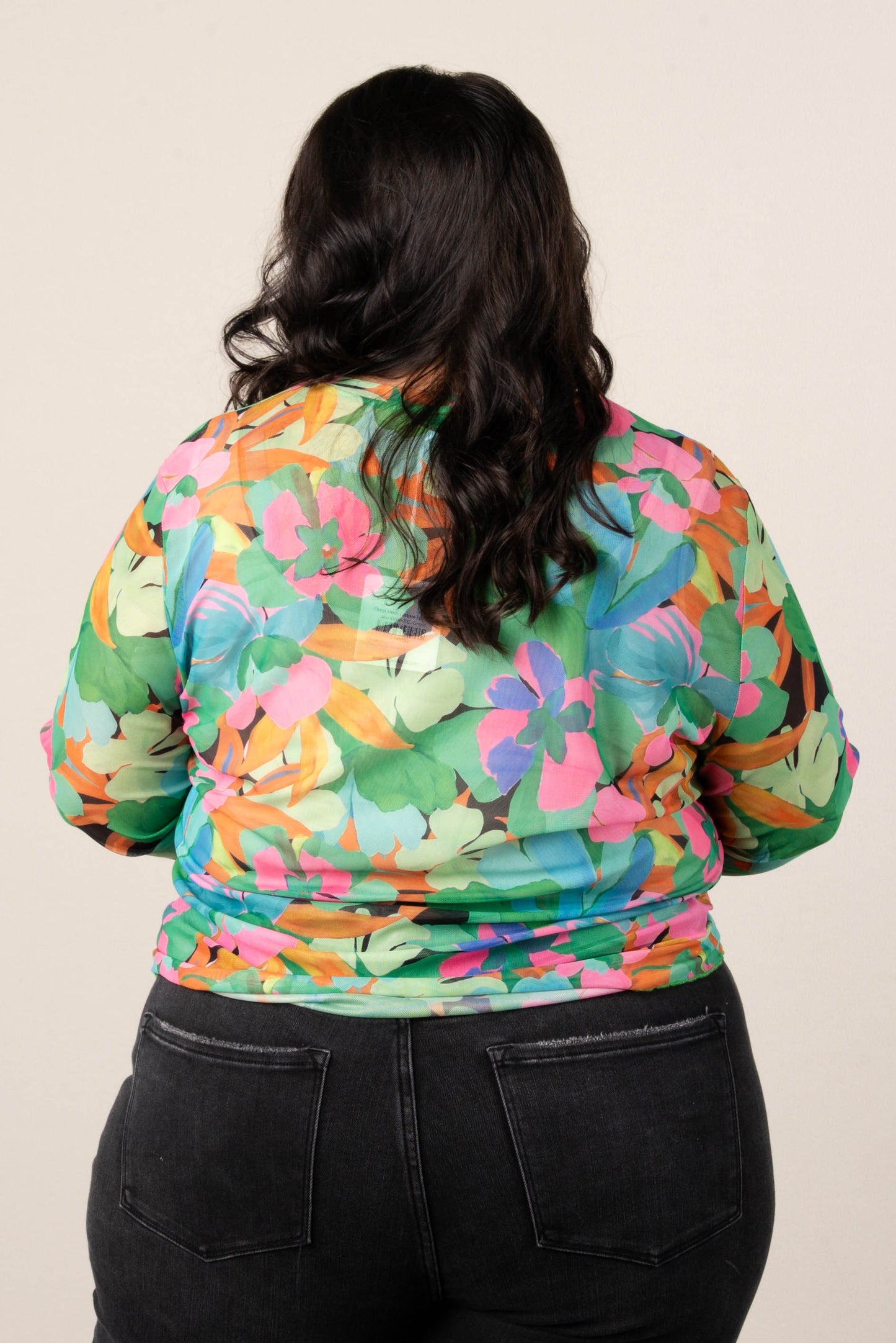 Back of plus size woman wearing a colorful floral mesh long sleeve top in green, pink, and orange tones, styled with high-waisted black jeans.