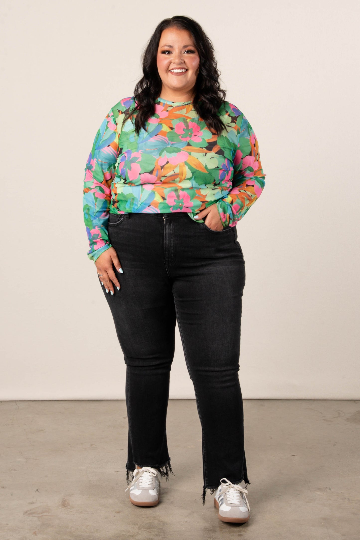 Smiling, plus size woman wearing a colorful floral mesh long sleeve top in green, pink, and orange tones, styled with high-waisted black jeans and tennis shoes.