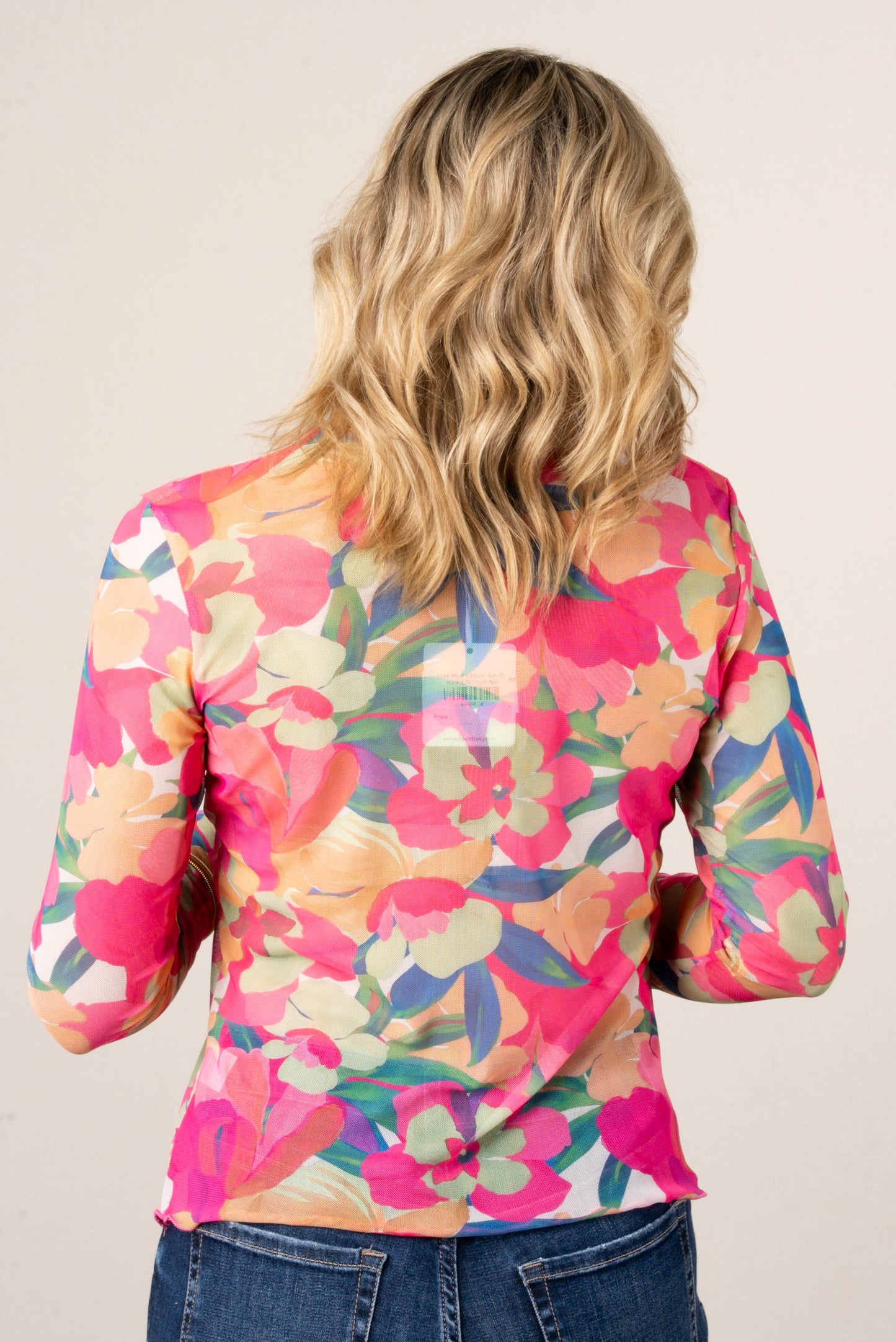 Back of a woman wearing a long sleeve floral mesh top in bright pink, orange, green, and cream tones, paired with blue jeans.