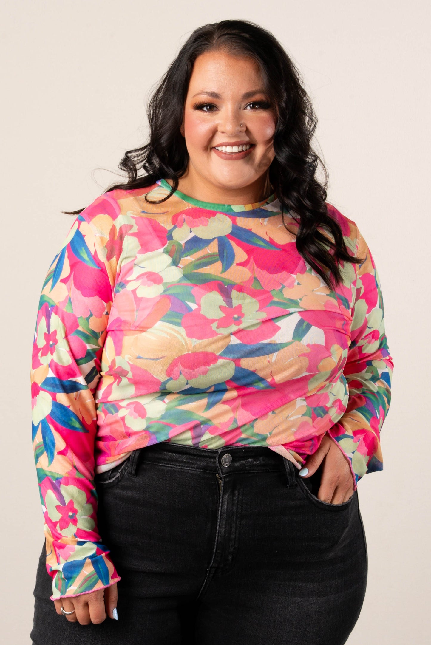 Smiling woman wearing a plus size long sleeve floral mesh top with vibrant green, pink, orange, and blue tropical flower print, paired with black jeans.