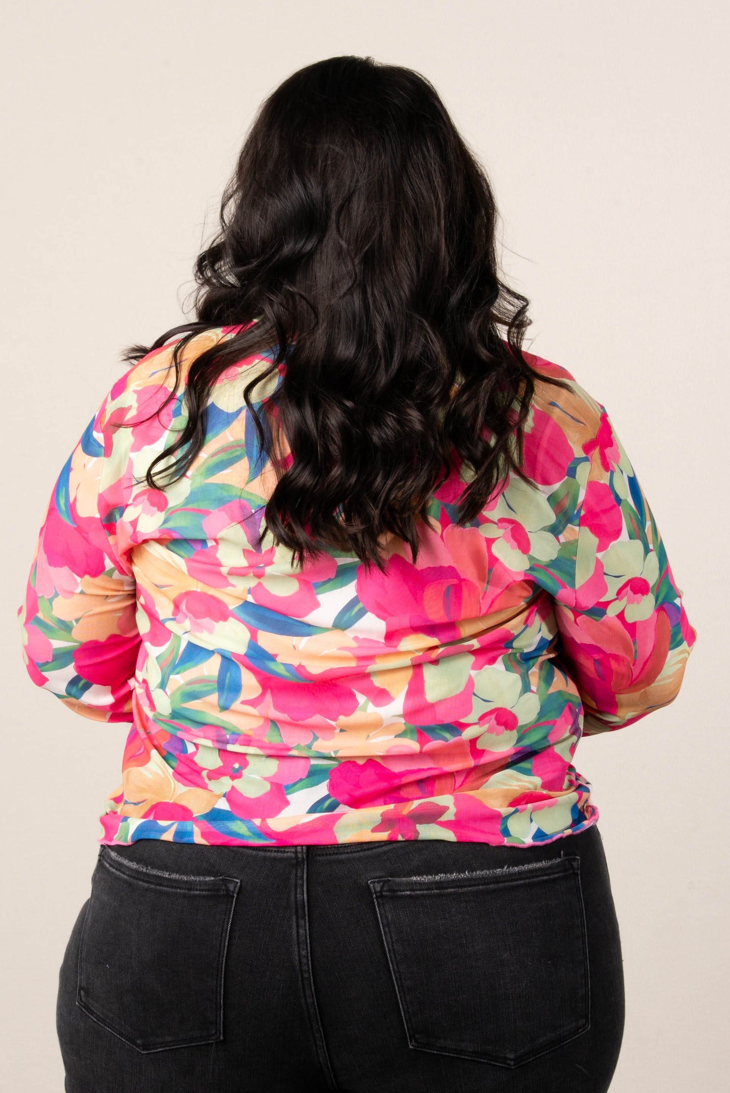 Back of a woman wearing a plus size long sleeve floral mesh top with vibrant green, pink, orange, and blue tropical flower print, paired with black jeans.