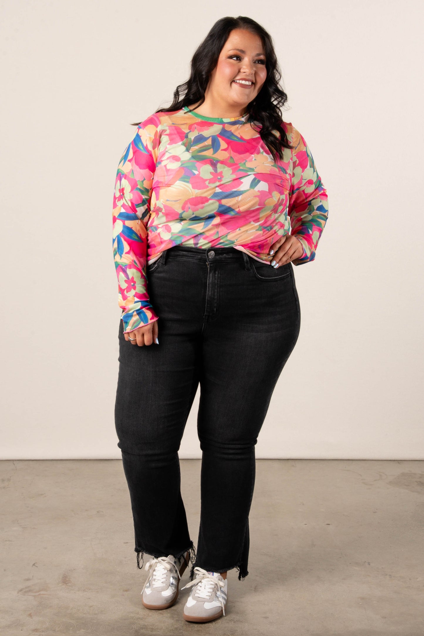 Smiling woman wearing a plus size long sleeve floral mesh top with vibrant green, pink, orange, and blue tropical flower print, paired with black jeans.