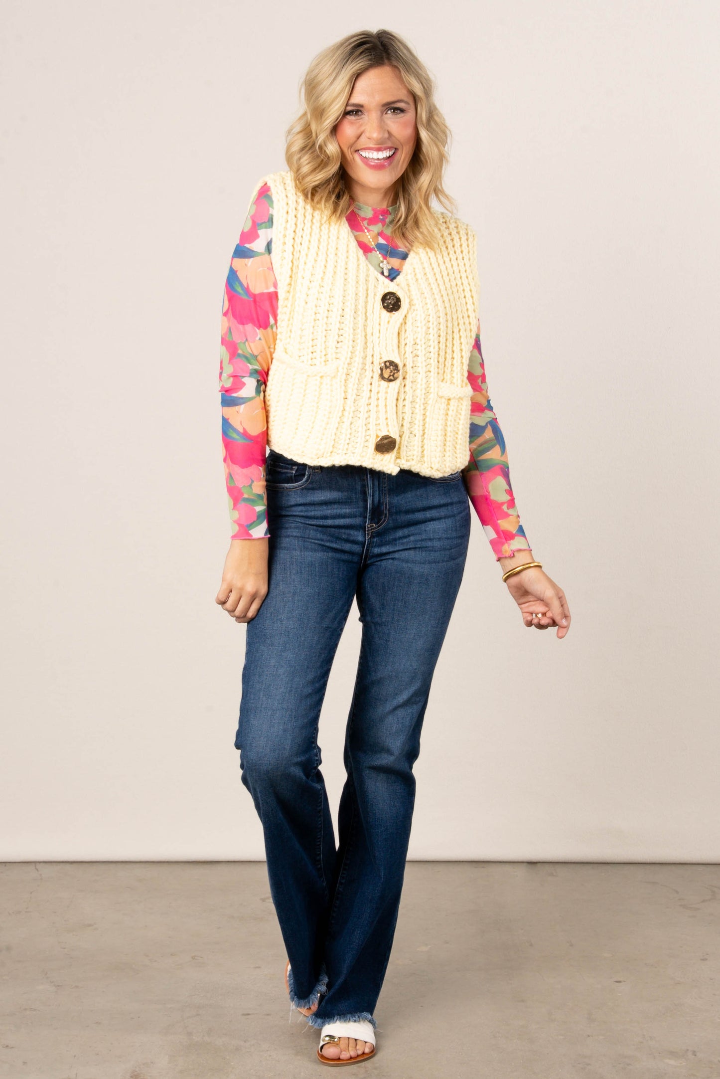 Smiling woman wearing a colorful pink floral mesh long sleeve top layered under a chunky yellow knit vest with large gold buttons, paired with dark wash bootcut jeans and open-toe sandals.