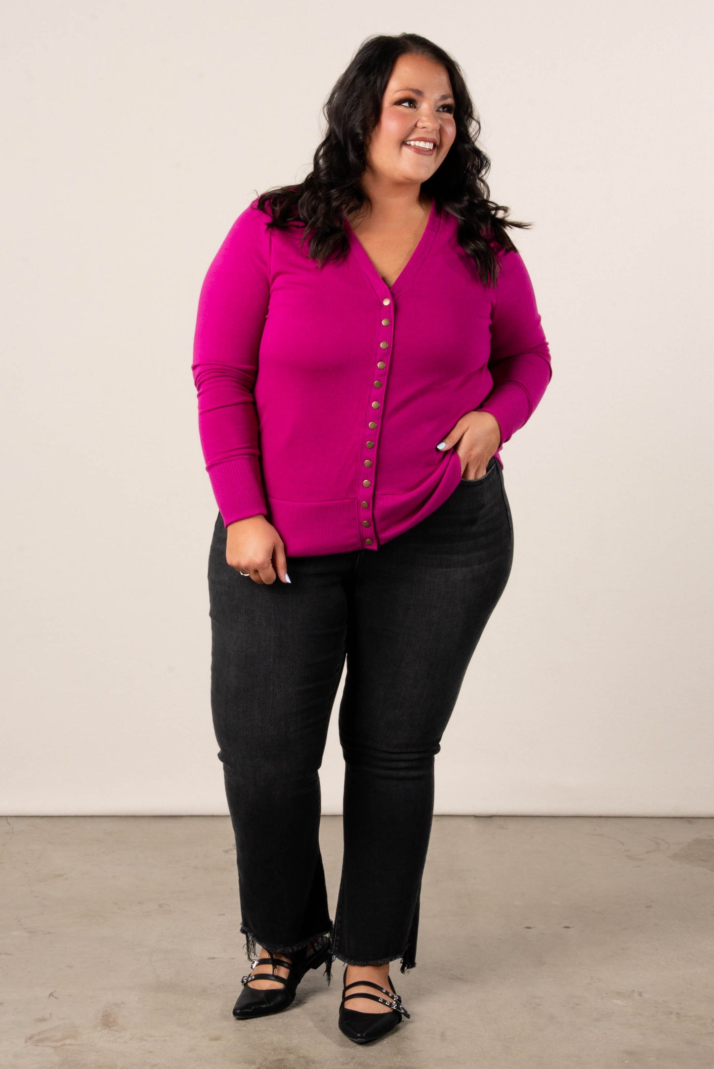 Smiling, plus size woman wearing a magenta button-front cardigan, styled with high-waisted black jeans and black pointed dress flats.