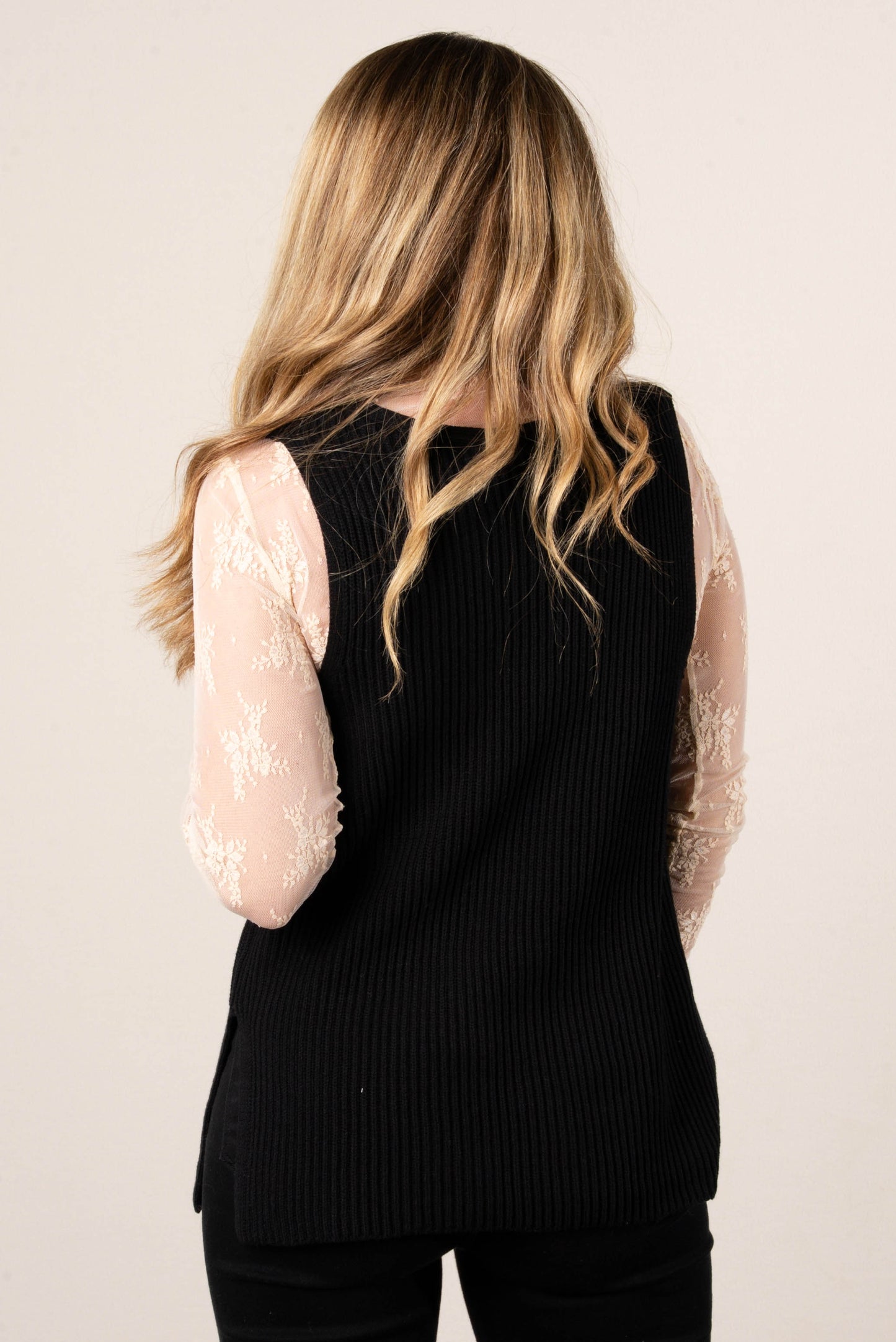 A back view of a woman with long, blonde hair. She is wearing a black ribbed sweater vest over a sheer beige lace top.