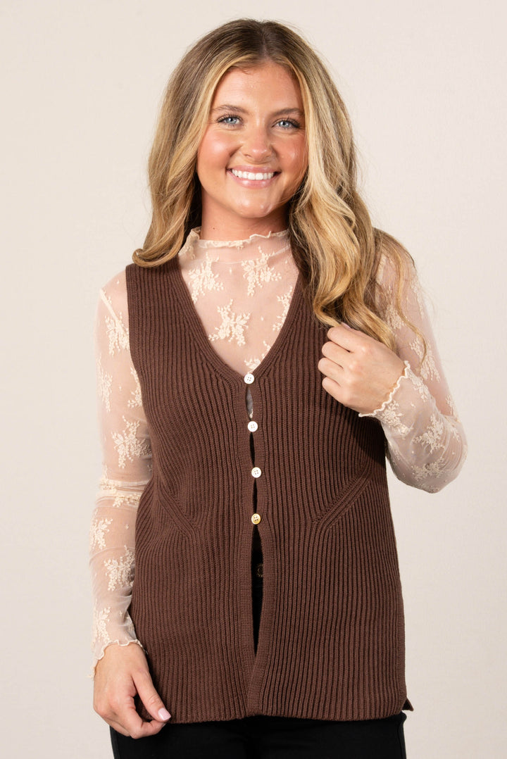 A close-up of a woman with long, blonde hair, smiling and looking directly at the camera. She is wearing a brown ribbed sweater vest over a sheer beige lace top. The sweater vest has a v-neckline and pearlescent buttons down the front.