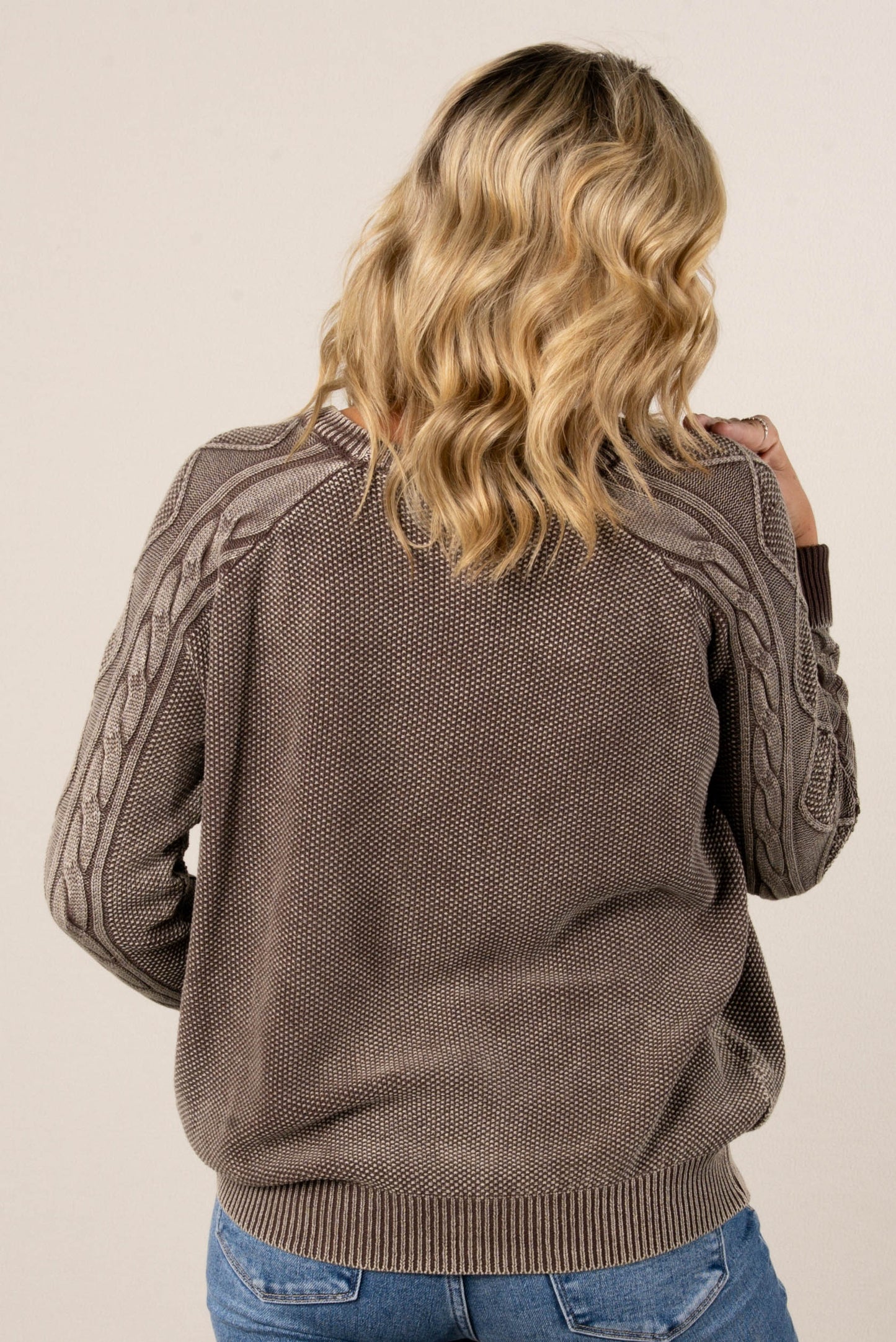 A back view of a woman with blonde hair, wearing a brown knit sweater with a textured knit on the back and cable-knit details on the sleeves. She's also wearing blue jeans.