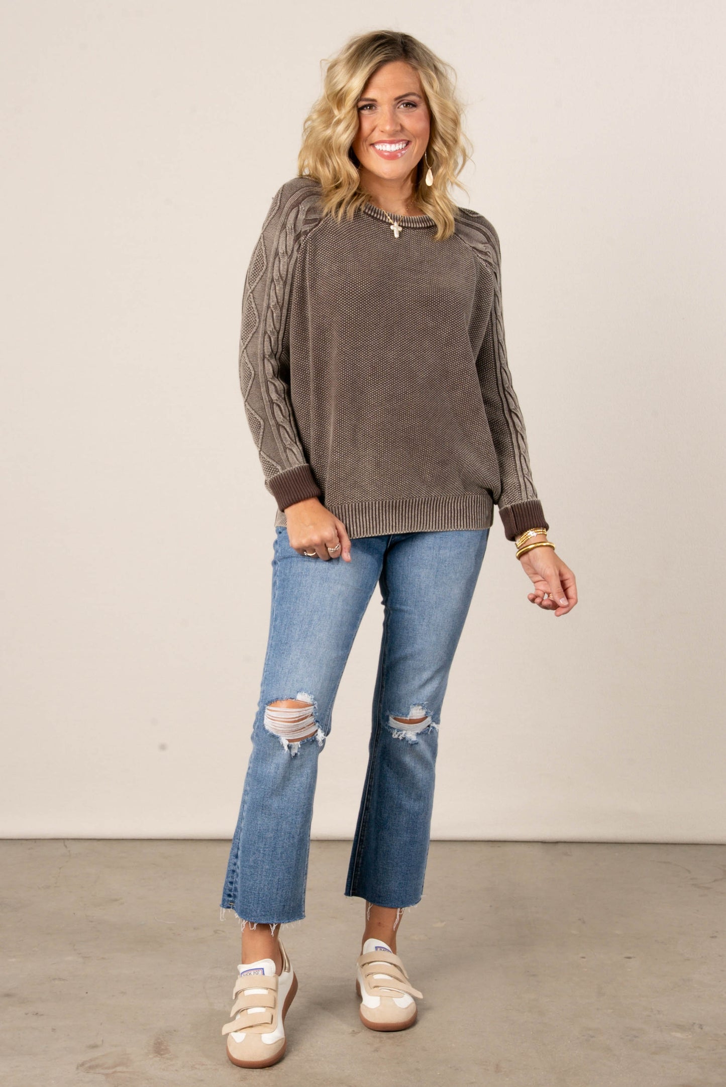A full-length photo of a woman with blonde hair, smiling at the camera. She's wearing a brown knit sweater with cable-knit details on the raglan sleeves. The sweater has a crew neckline and a ribbed hem and cuffs. She is also wearing distressed jeans and cream-colored sneakers with velcro straps.
