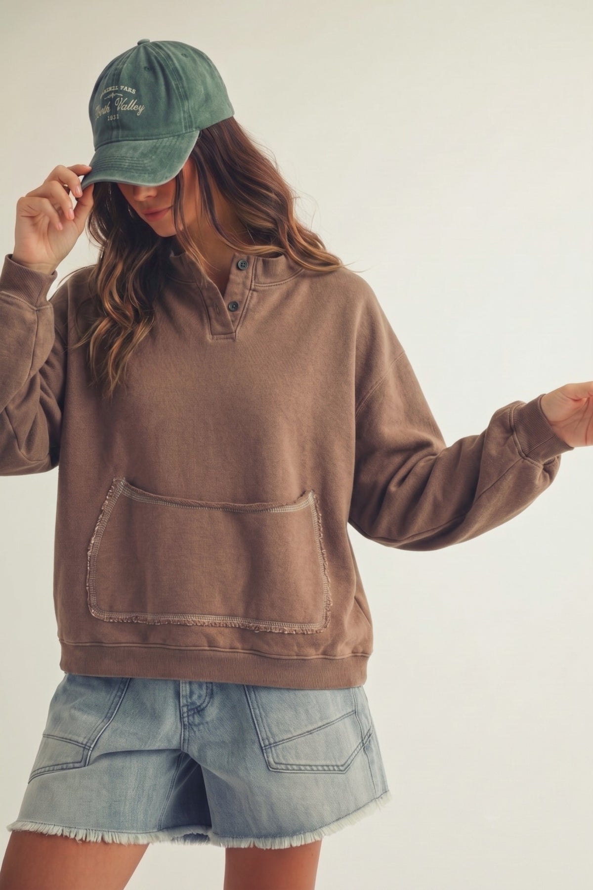 Woman wearing a brown henley pouch pocket pullover with denim shorts and a green cap