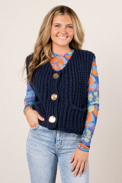 Smiling woman wearing a colorful blue floral mesh long sleeve top layered under a chunky navy knit vest with large gold buttons, paired with light wash jeans.