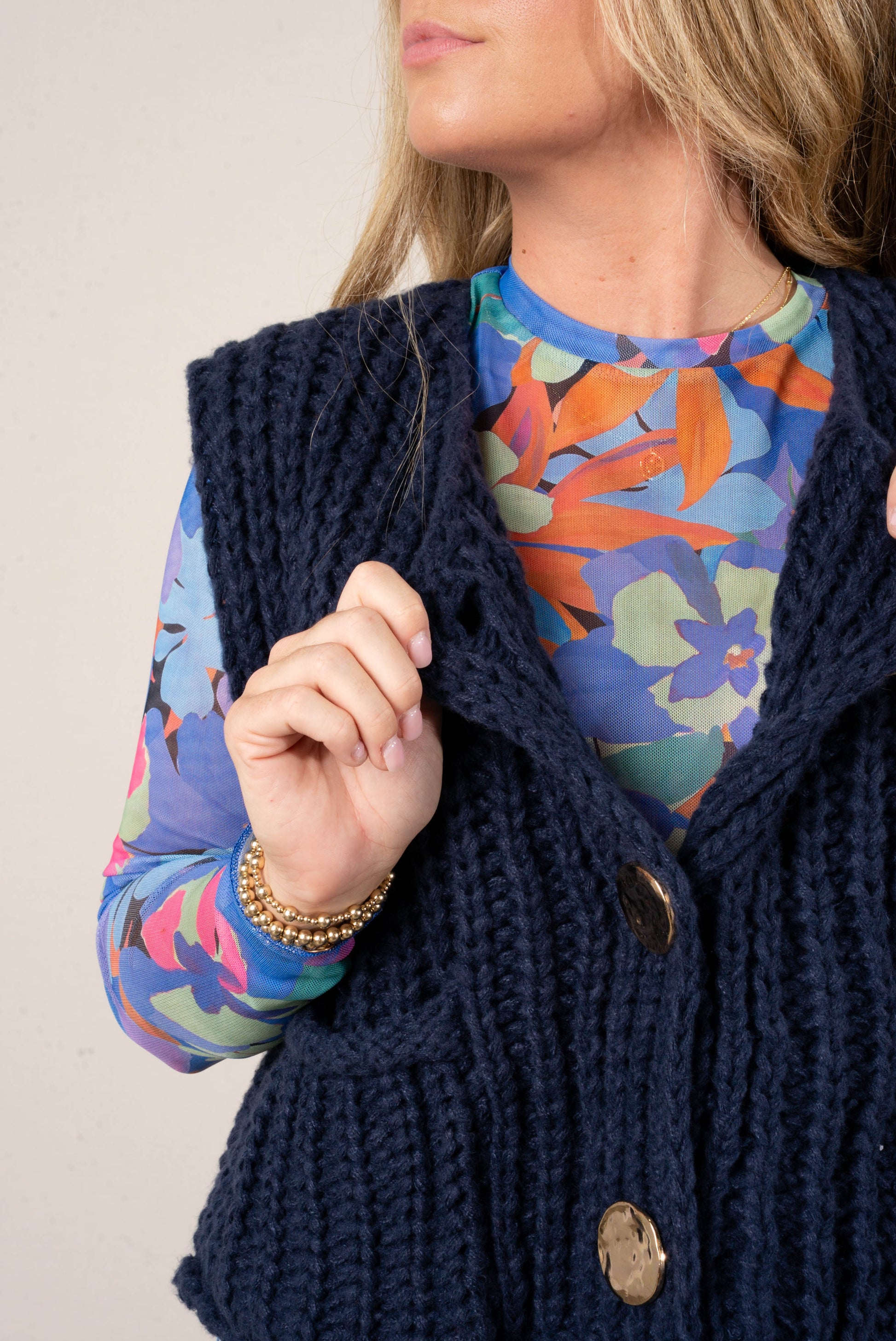 Close up of a woman wearing a bright blue floral print mesh top layered under a navy, chunky knit sweater vest with large gold buttons.