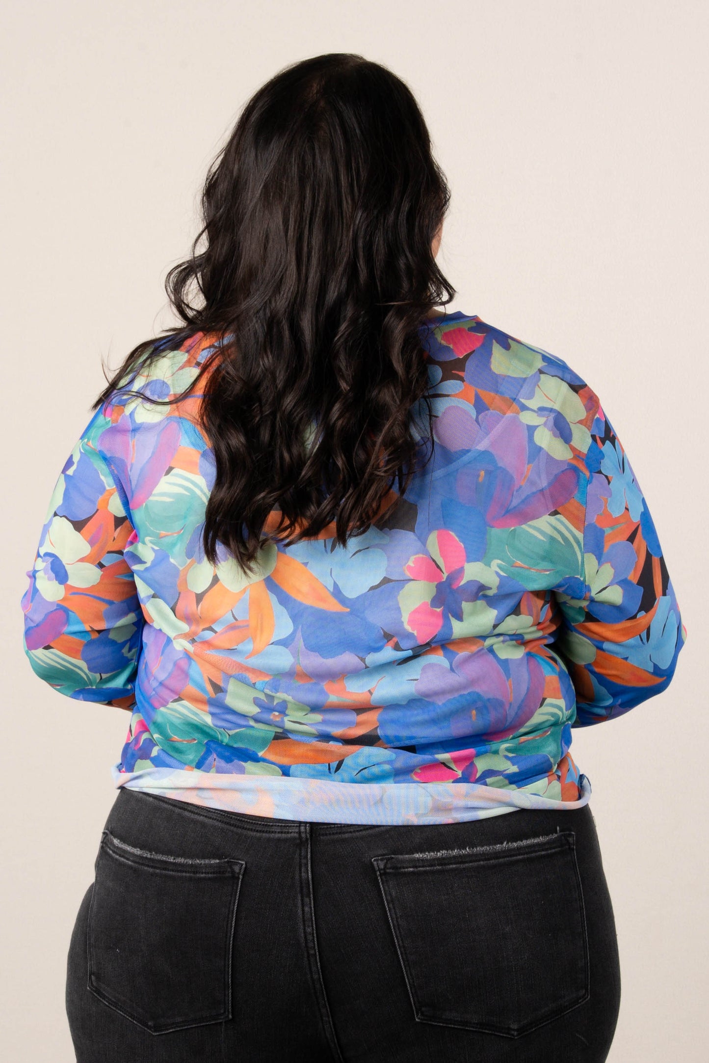 Back of a woman wearing a colorful floral mesh long sleeve top in blue, orange, pink, and green tones, paired with black high-waisted jeans.