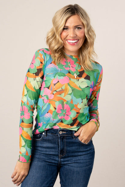 Smiling woman wearing a colorful floral mesh long sleeve top in green, pink, and orange tones, styled with high-waisted blue jeans.