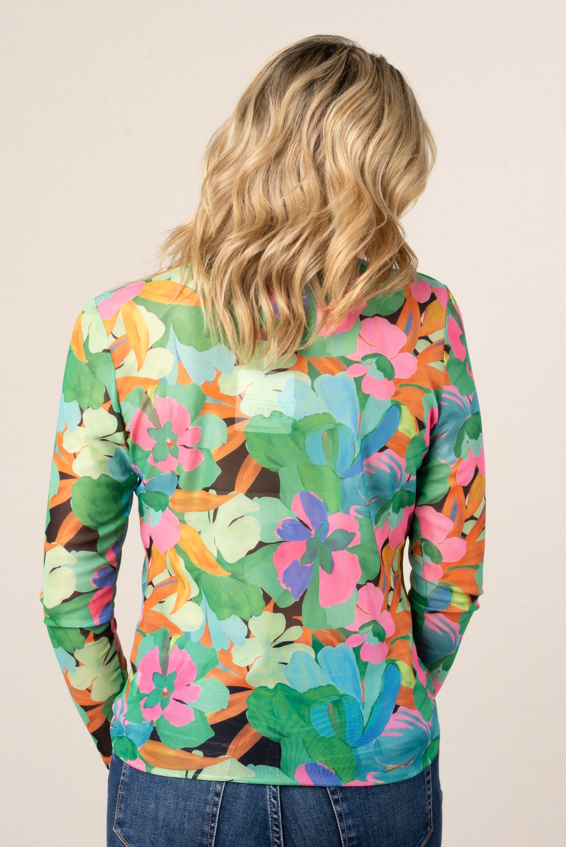 Back of a woman wearing a colorful floral mesh long sleeve top in green, pink, and orange tones, styled with high-waisted blue jeans.