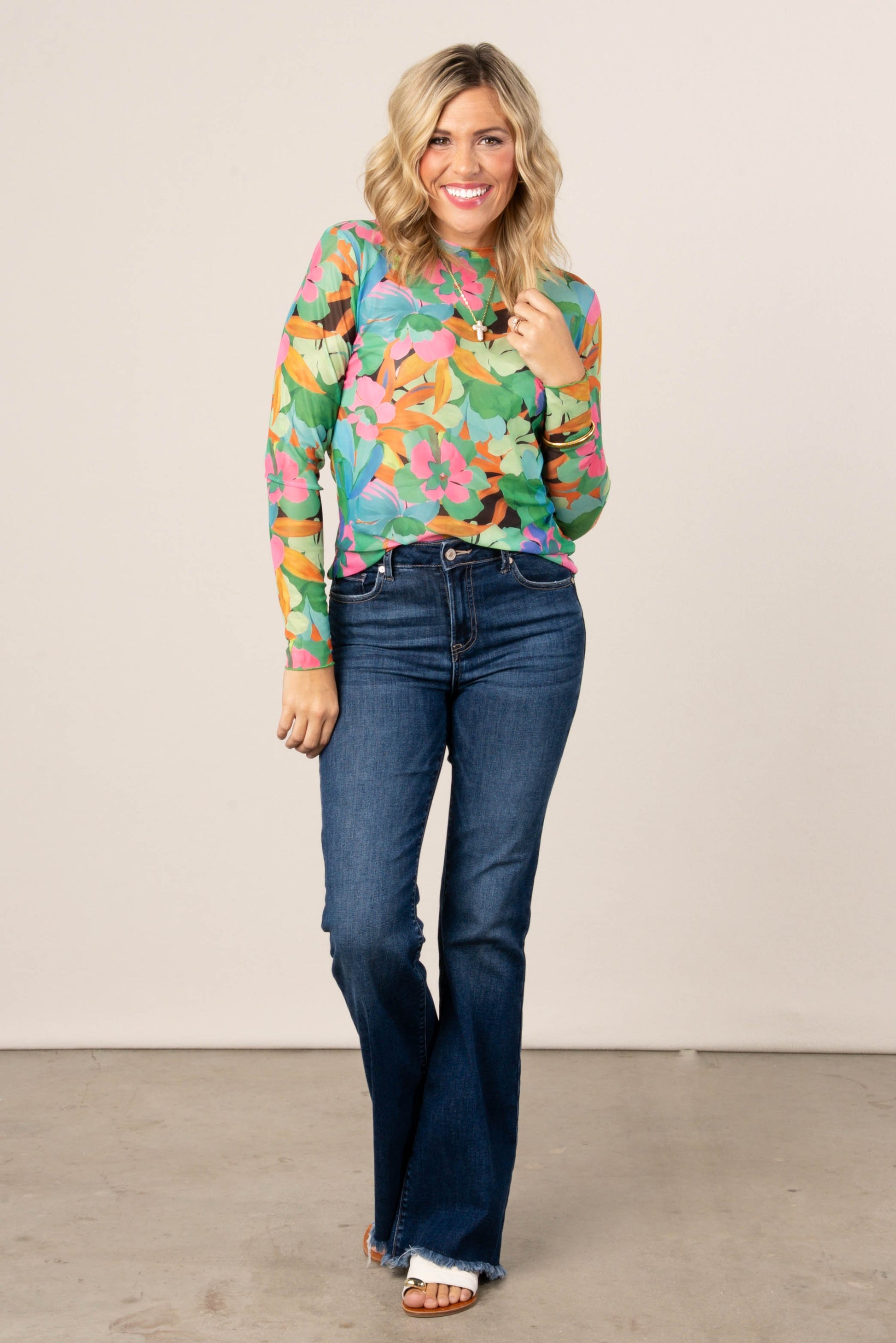 Smiling woman wearing a colorful floral mesh long sleeve top in green, pink, and orange tones, styled with high-waisted blue flare jeans and sandals.