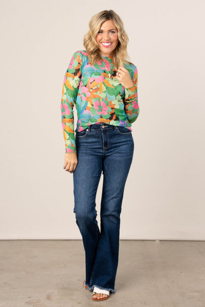 Smiling woman wearing a colorful floral mesh long sleeve top in green, pink, and orange tones, styled with high-waisted blue flare jeans and sandals.