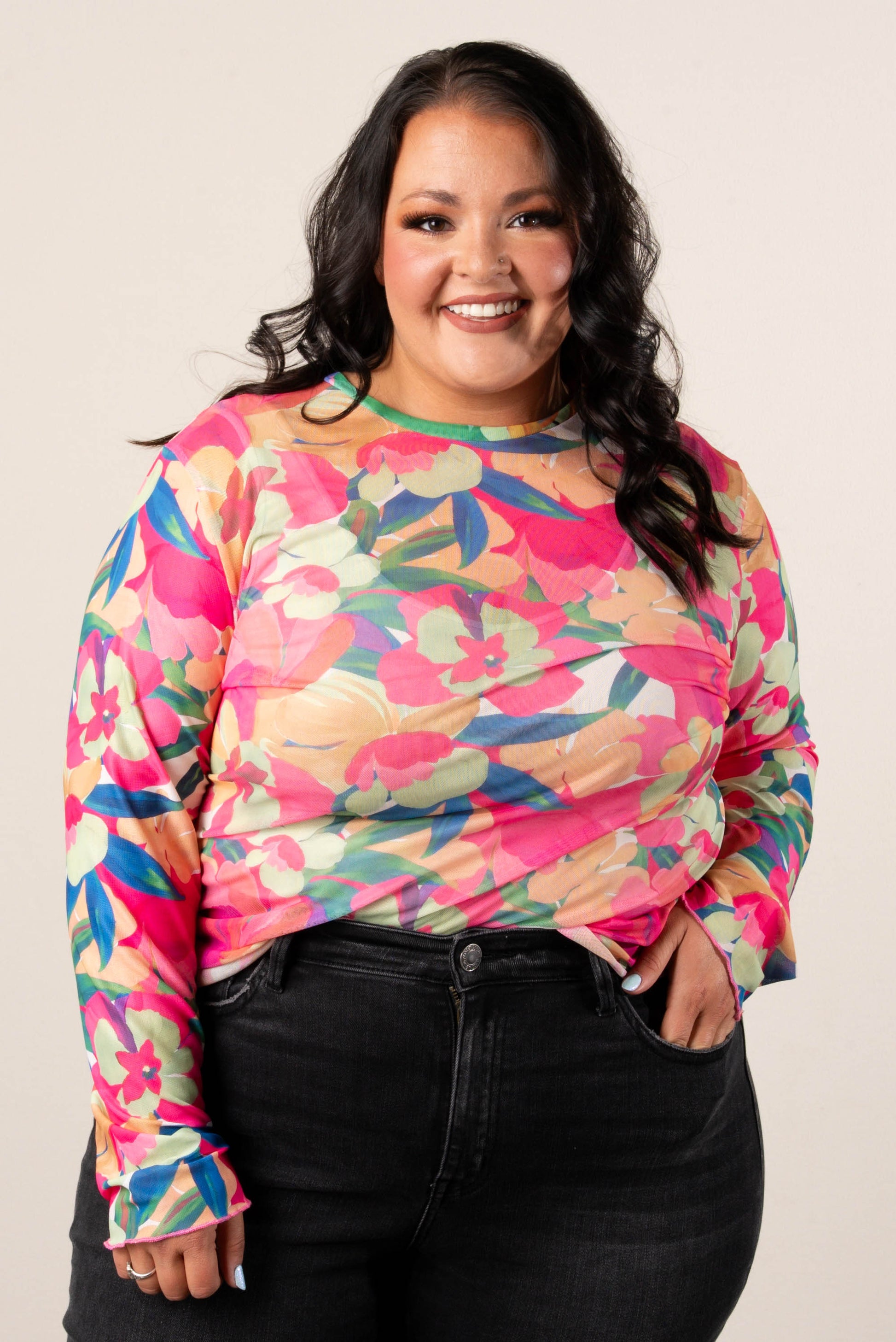 Smiling woman wearing a plus size long sleeve floral mesh top with vibrant green, pink, orange, and blue tropical flower print, paired with black jeans.