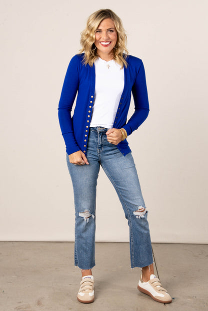 Smiling woman wearing a cobalt blue button-front cardigan over a white t-shirt, styled with cropped,  high-waisted light wash jeans and tennis shoes