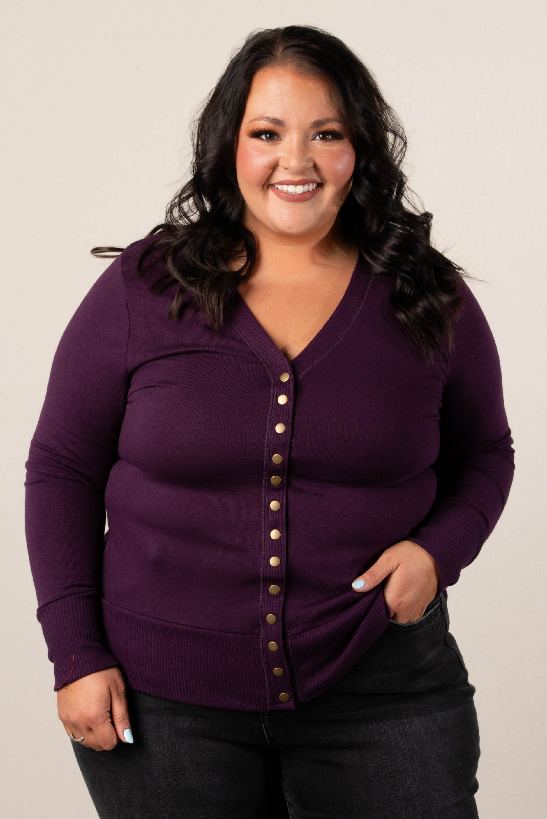 Smiling plus size woman wearing an eggplant colored, button-front cardigan, styled with high-waisted black jeans.