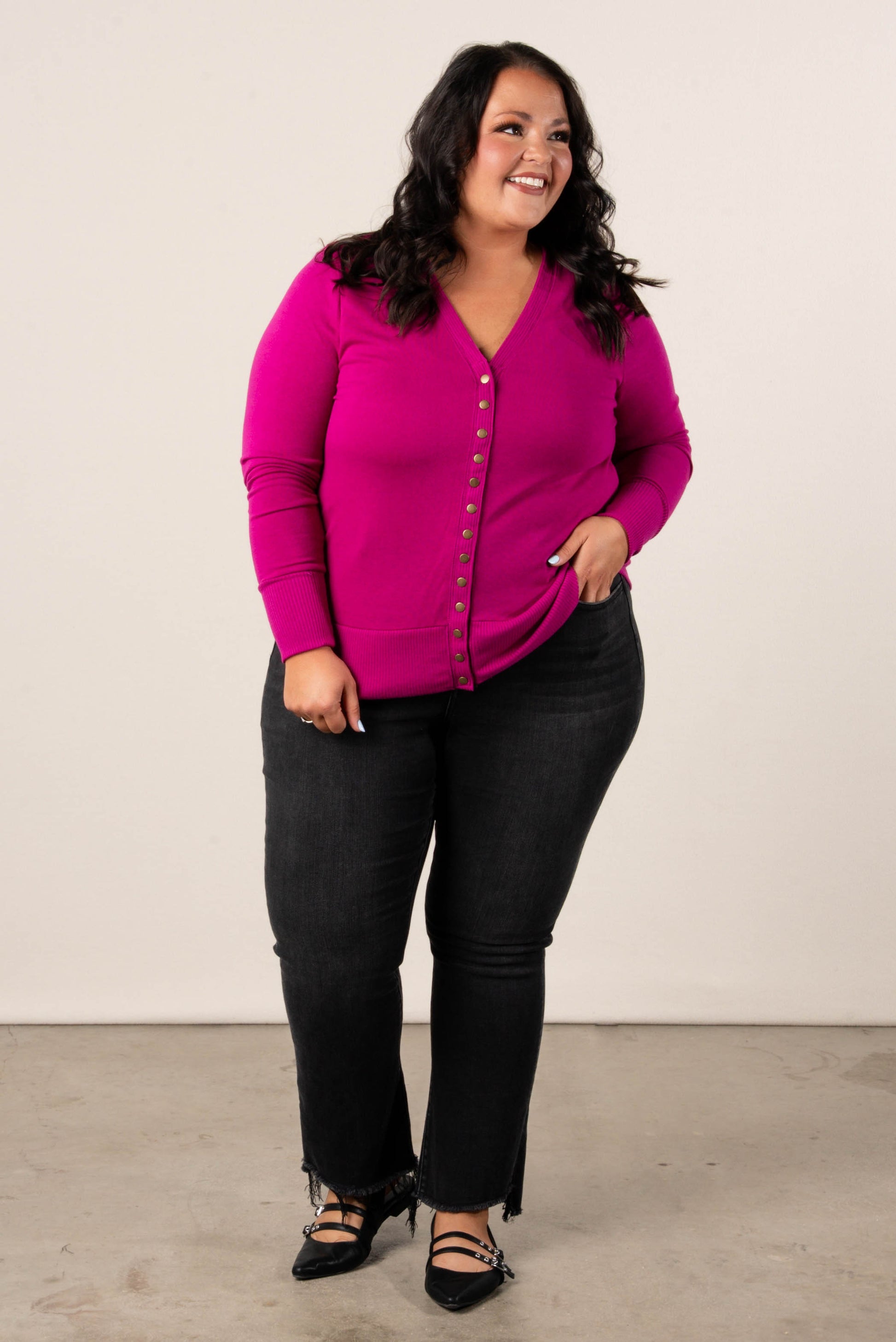 Smiling, plus size woman wearing a magenta button-front cardigan, styled with  high-waisted black jeans and black pointed dress flats.