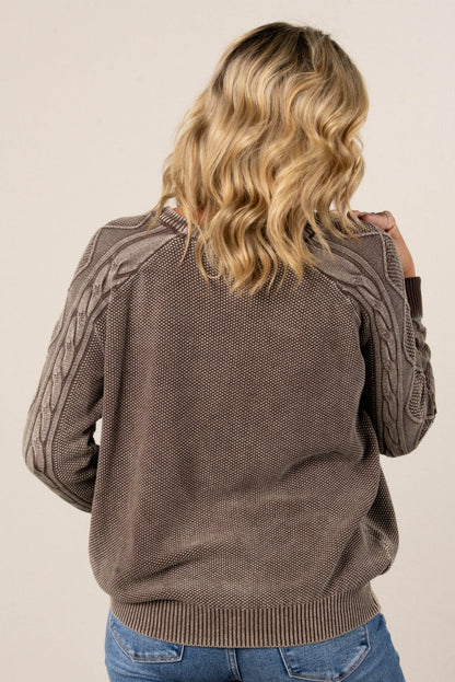 A back view of a woman with blonde hair, wearing a brown knit sweater with a textured knit on the back and cable-knit details on the sleeves. She's also wearing blue jeans.