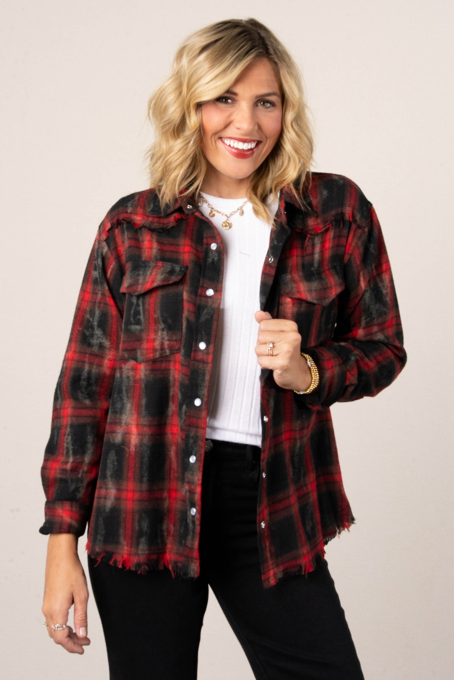 A woman with blonde hair smiling while wearing an open red and black distressed plaid button-down shirt. The shirt has two front pockets with buttons. She is wearing it over a white top and black jeans.