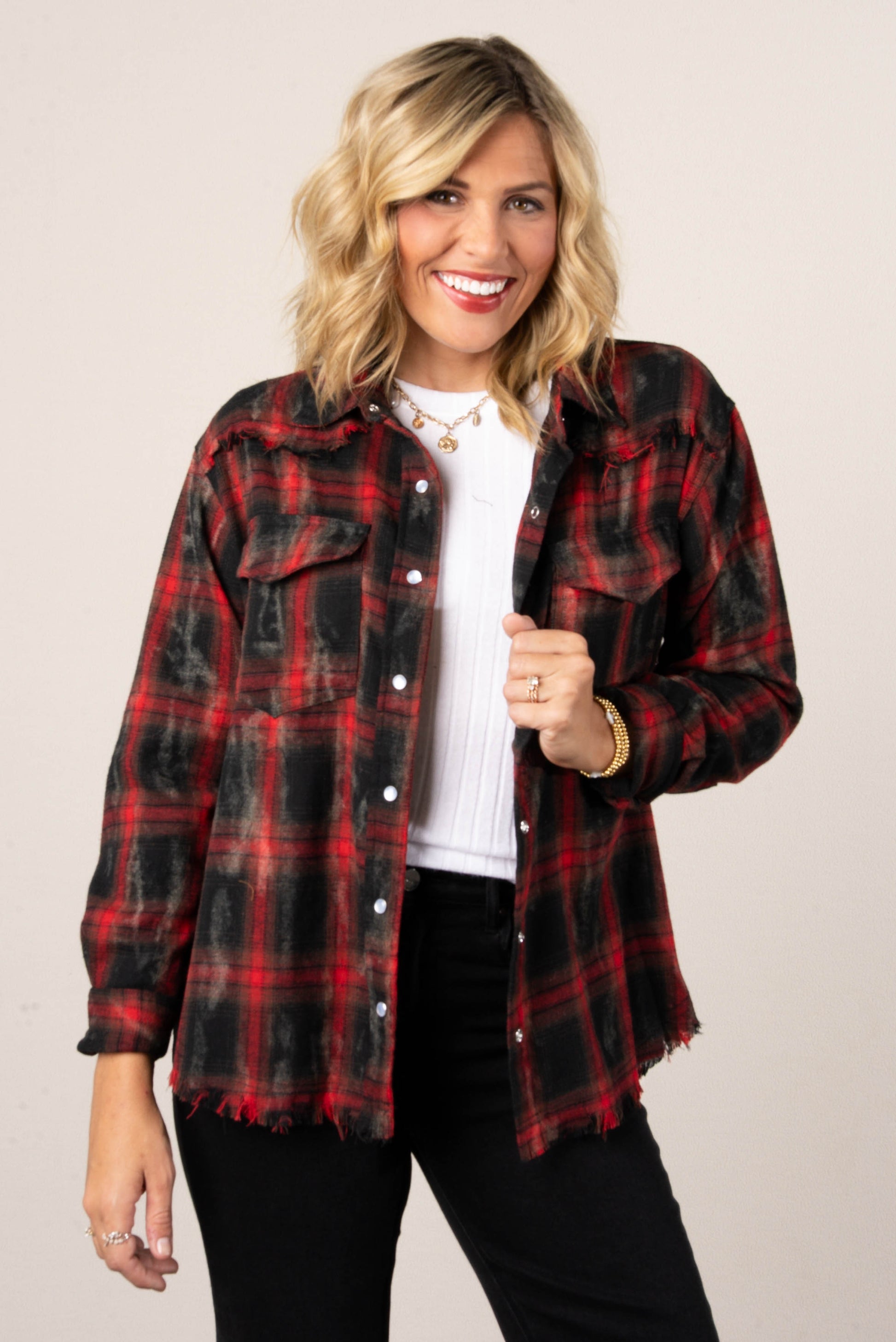 A woman with blonde hair smiling while wearing an open red and black distressed plaid button-down shirt. The shirt has two front pockets with buttons. She is wearing it over a white top and black jeans.