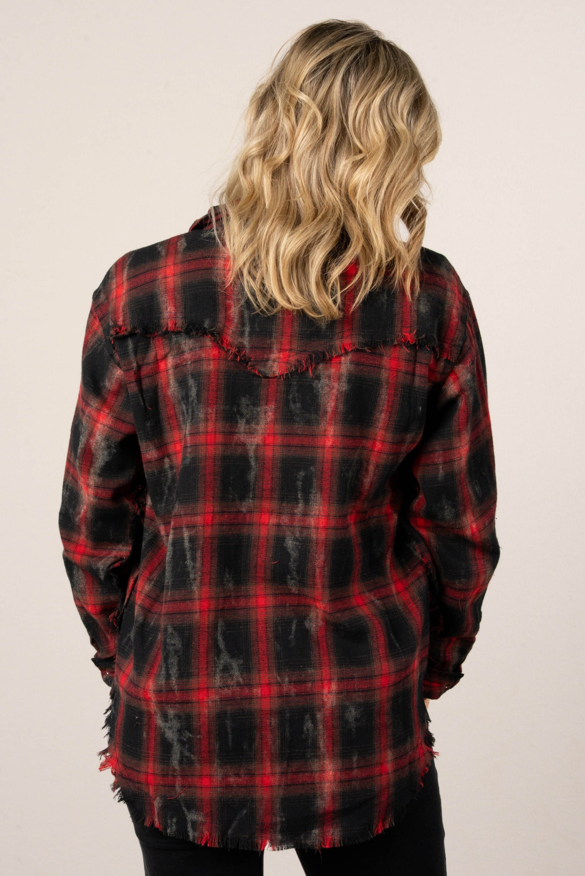 A back view of a woman with blonde hair, wearing a red and black distressed plaid button-down shirt. The back of the shirt features a yoke with a frayed edge.