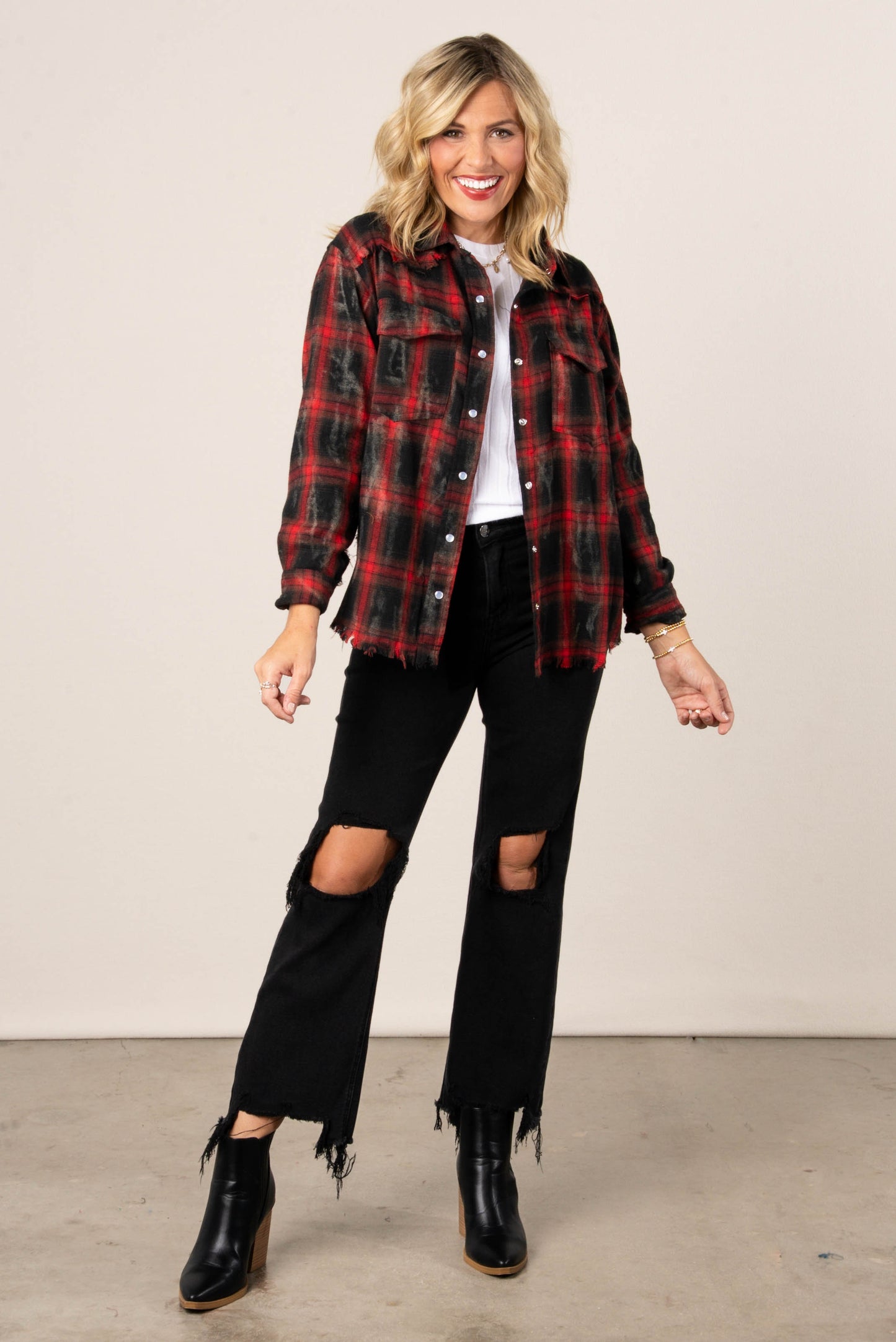 A full-length photo of a woman with blonde hair, wearing an open red and black distressed plaid button-down shirt over a white top. She is also wearing distressed black jeans with a cropped, frayed hem and black pointed-toe booties.