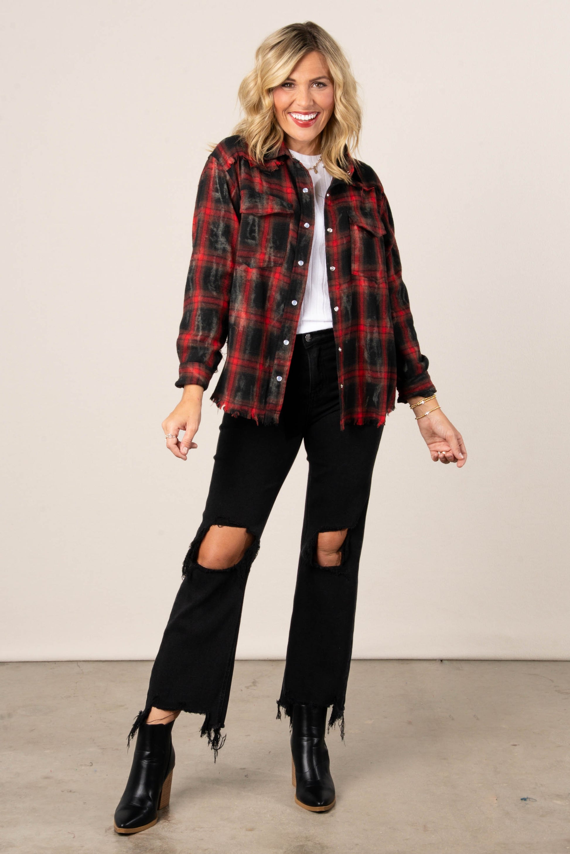 A full-length photo of a woman with blonde hair, wearing an open red and black distressed plaid button-down shirt over a white top. She is also wearing distressed black jeans with a cropped, frayed hem and black pointed-toe booties.