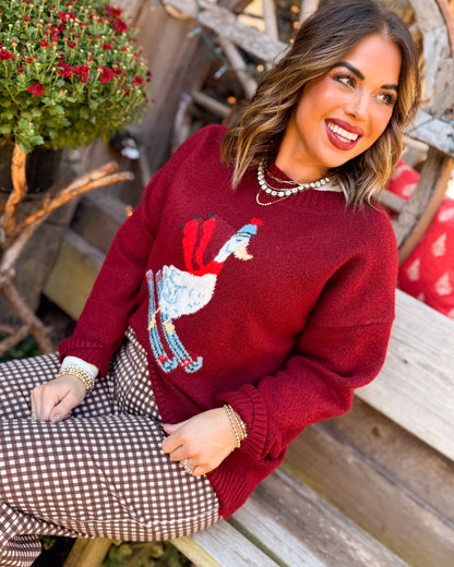 Woman wearing a red skiing duck knit sweater styled with brown houndstooth pants seated on a bench
